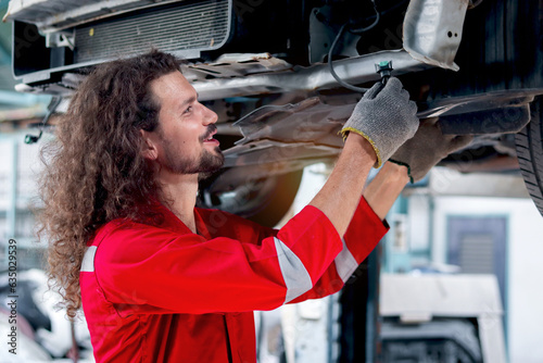 Long hair mechanic man in red uniform work with lifted vehicle, inspect car engine system underbody, auto mechanic check wheel at garage, technician repairing customer car automobile at repair service
