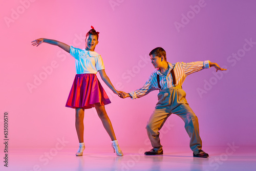 Talented, artisitc children, boy and girl in retro clothes dancing lindy hop against pink studio background in neon light. Concept of childhood, hobby, active lifestyle, performance, art, fashion