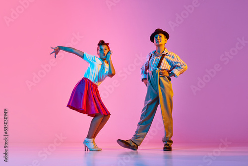 Emotional, beautiful children, boy and girl in retro costumes dancing lindy hop against pink studio background in neon light. Concept of childhood, hobby, active lifestyle, performance, art, fashion