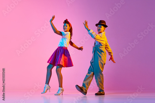 Emotional and stylish children, boy and girl in retro clothes dancing lindy hop against pink studio background in neon light. Concept of childhood, hobby, active lifestyle, performance, art, fashion