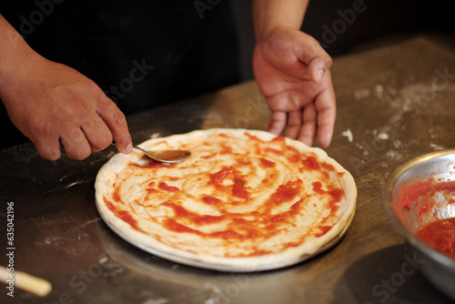 Closeup image of pizzamaker spreading tomato sauce on pizza dough