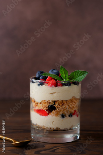 Sweet dessert in a glasses with berries, mint, whipped coconut cream and biscuit on a brown background. Healthy food, vegan, sugar, gluten and lactose free.