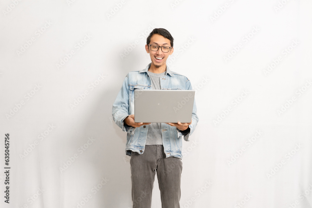 A confident male model gives happy expression when he looking to his laptop. Indonesian or southeast asian model isolated with white wall background. A confident male model gives happy expression when he looking to his laptop. Indonesian or southeast asian model isolated with white wall background.