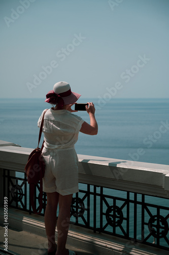 giovane donna con cappello e nastro ammira il mare dall'alto scattando foto dal suo smartphone in un contesto di serenità e pace photo