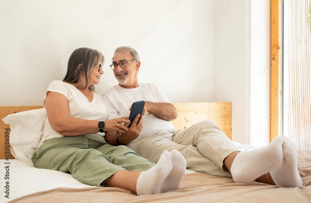 Smiling senior european man and woman watch video on phone, sit on bed in bedroom interior