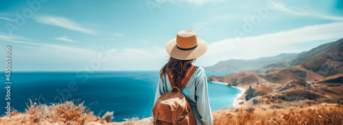 A woman standing on a cliff overlooking the ocean and the coastline. The woman is wearing a straw hat, a blue sweater and carrying a brown backpack. Warm and peaceful mood.