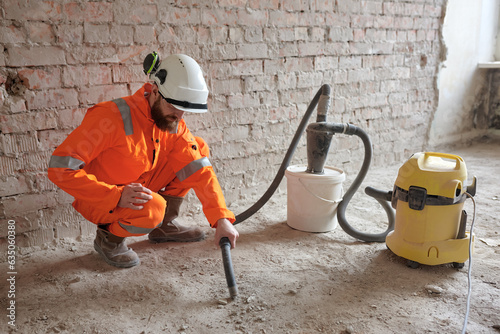 Male constriction site worker cleaning concrete floor from dust with vacuum cleaner after demolishing jobs.