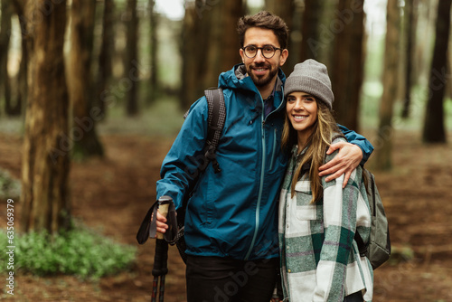 Positive young caucasian couple in jackets with trekking sticks walk in forest together, enjoy trip