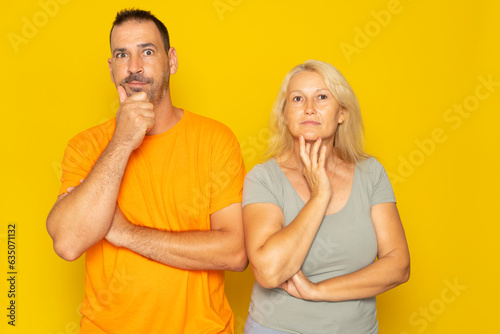 Caucasian real couple in thoughtful pose with hand on chin making a difficult decision, isolated on yellow studio background.