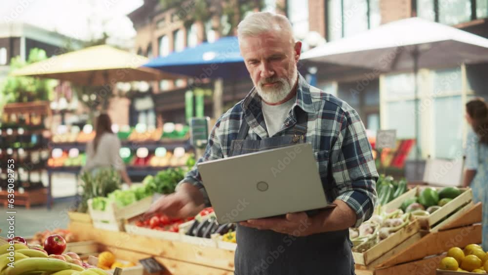 Senior Farmers Market Manager Using Laptop Computer while Standing Next to Street Vendor Booths with Fresh Organic Agricultural Products. Adult Male Managing Business Development in Rural Community Senior Farmers Market Manager Using Laptop Computer while Standing Next to Street Vendor Booths with Fresh Organic Agricultural Products. Adult Male Managing Business Development in Rural Community