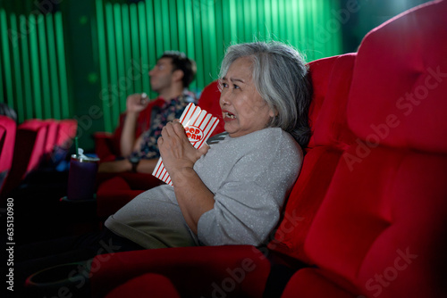 Asian senior woman hold popcorn and look scary during watch movie in cinema theater and Caucasian man sit near her on the same roll of seats.