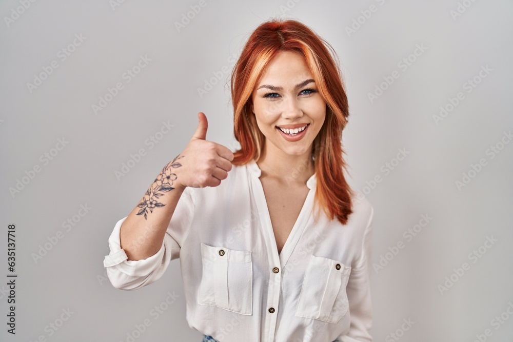 Young caucasian woman standing over isolated background doing happy thumbs up gesture with hand. approving expression looking at the camera showing success. Young caucasian woman standing over isolated background doing happy thumbs up gesture with hand. approving expression looking at the camera showing success.