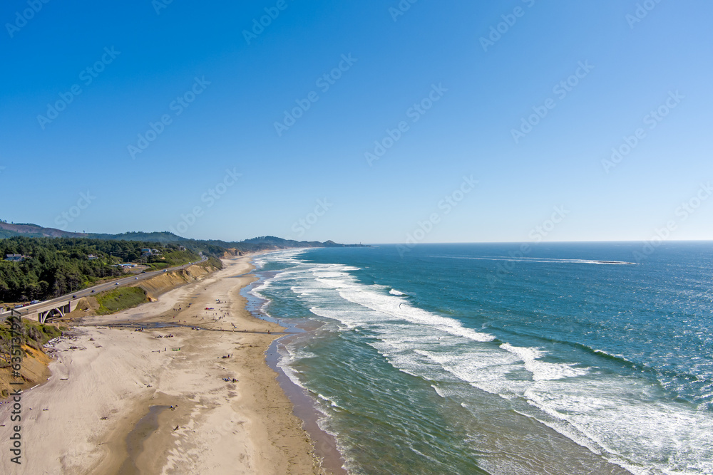 Aerial view of Beverly Beach on the Oregon Coast with gentle breaking waves on the sandy beach, the Yaquina Head Lighthouse and beach goers in the distance and a bright blue sky with copy space.