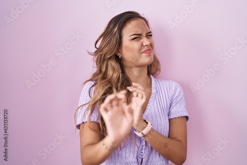 Young hispanic woman standing over pink background disgusted expression, displeased and fearful doing disgust face because aversion reaction. photo