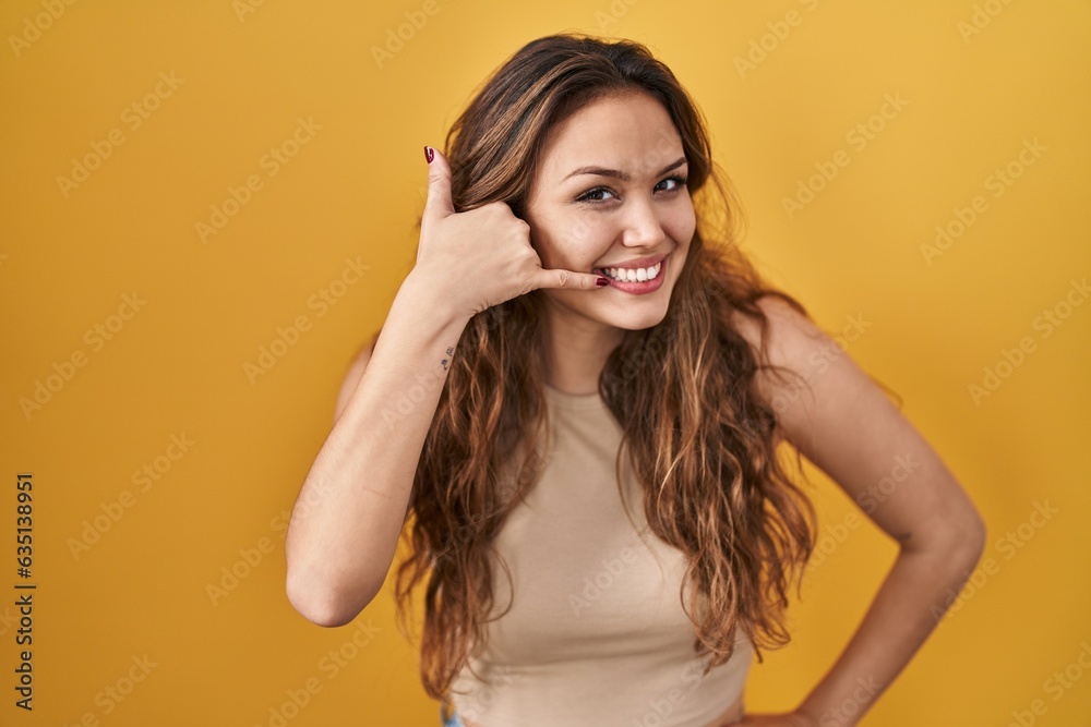 Young hispanic woman standing over yellow background smiling doing phone gesture with hand and fingers like talking on the telephone. communicating concepts. Young hispanic woman standing over yellow background smiling doing phone gesture with hand and fingers like talking on the telephone. communicating concepts.