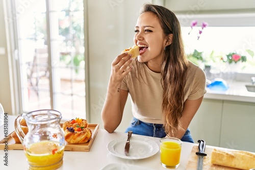Young beautiful hispanic woman eating bread with jam having breakfast at the kitchen