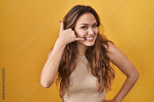 Young hispanic woman standing over yellow background smiling doing phone gesture with hand and fingers like talking on the telephone. communicating concepts.