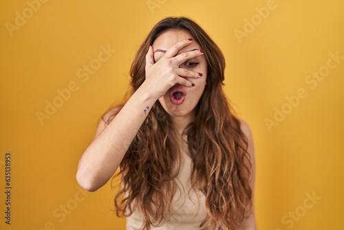 Young hispanic woman standing over yellow background peeking in shock covering face and eyes with hand, looking through fingers with embarrassed expression.