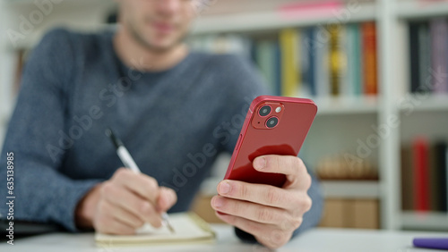 Young hispanic man student using smartphone writing on notebook at library university