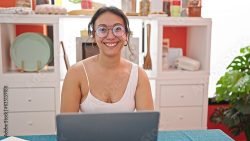 Young beautiful hispanic woman using laptop sitting on table smiling at dinning room