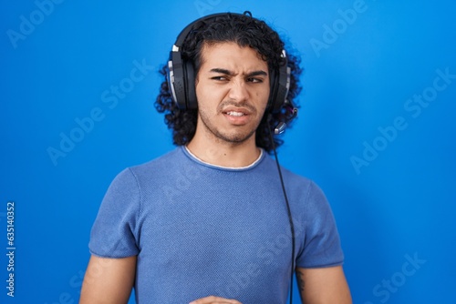 Hispanic man with curly hair listening to music using headphones disgusted expression, displeased and fearful doing disgust face because aversion reaction. with hands raised