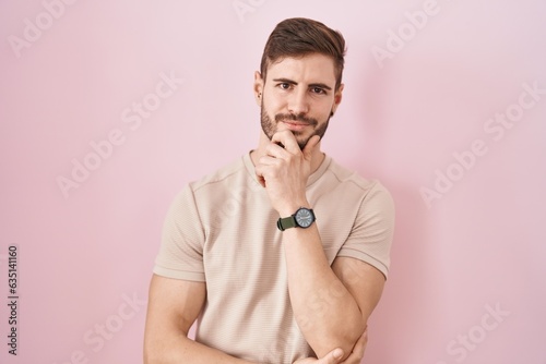 Hispanic man with beard standing over pink background looking confident at the camera smiling with crossed arms and hand raised on chin. thinking positive.