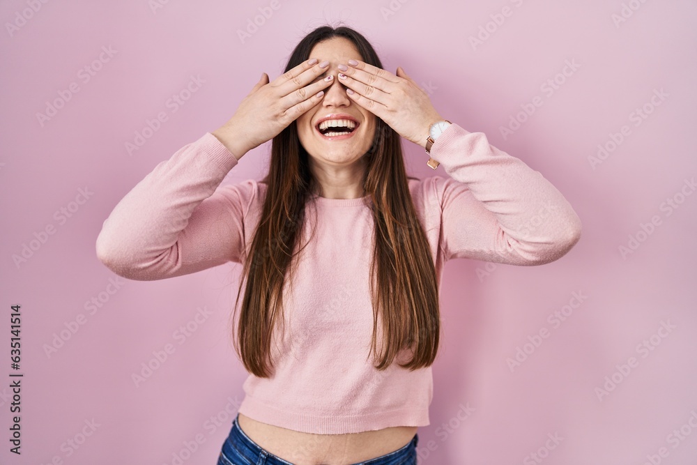 Young brunette woman standing over pink background covering eyes with hands smiling cheerful and funny. blind concept. Young brunette woman standing over pink background covering eyes with hands smiling cheerful and funny. blind concept.