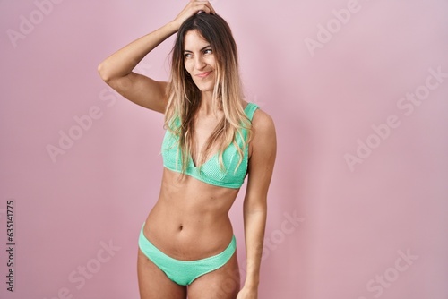 Young hispanic woman wearing bikini over pink background confuse and wondering about question. uncertain with doubt, thinking with hand on head. pensive concept.
