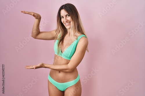 Young hispanic woman wearing bikini over pink background gesturing with hands showing big and large size sign, measure symbol. smiling looking at the camera. measuring concept. © Krakenimages.com