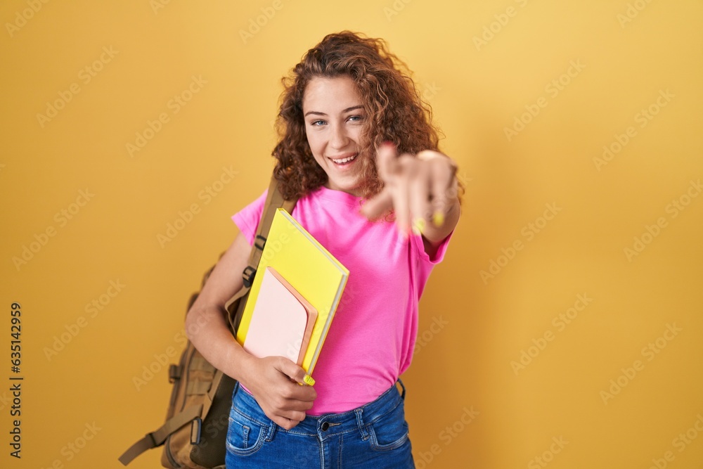 Young caucasian woman wearing student backpack and holding books pointing to you and the camera with fingers, smiling positive and cheerful Young caucasian woman wearing student backpack and holding books pointing to you and the camera with fingers, smiling positive and cheerful
