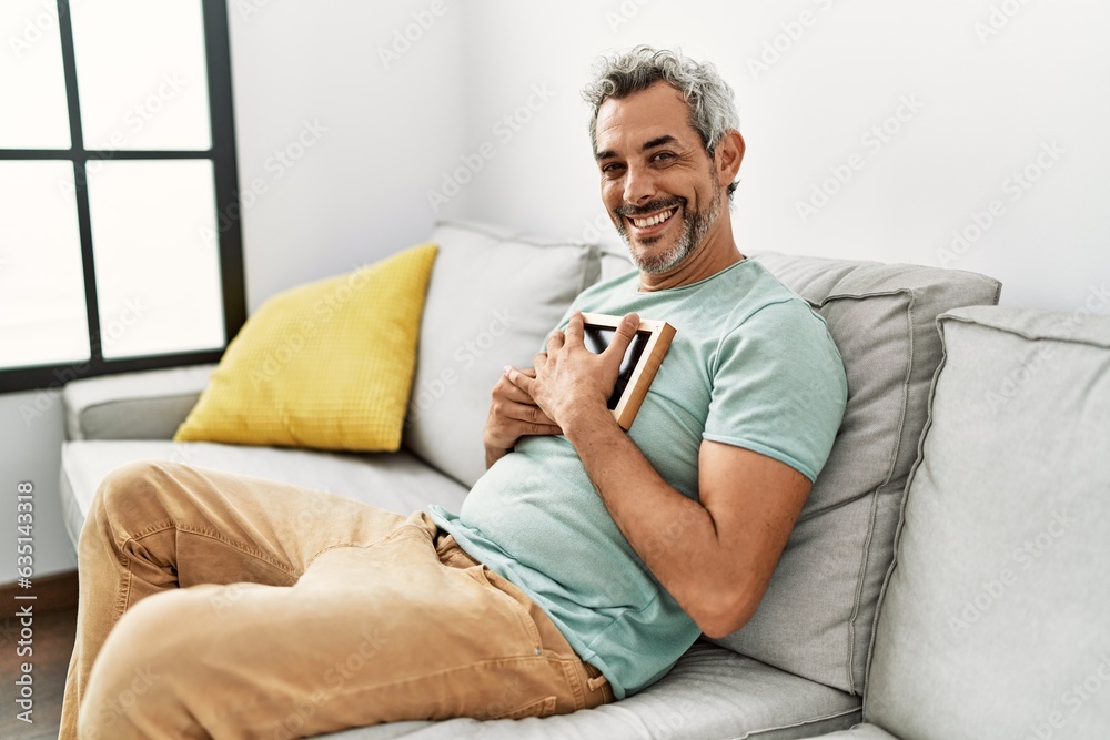 Middle age grey-haired man hugging picture sitting on sofa at home