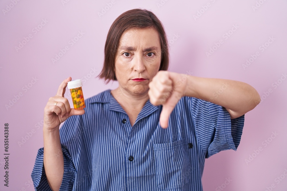 Middle age hispanic woman holding pills with angry face, negative sign showing dislike with thumbs down, rejection concept Middle age hispanic woman holding pills with angry face, negative sign showing dislike with thumbs down, rejection concept