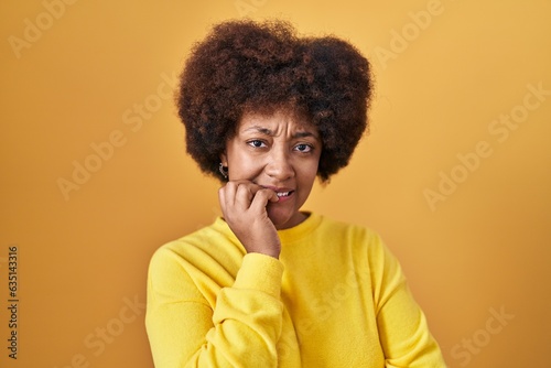 Young african american woman standing over yellow background looking stressed and nervous with hands on mouth biting nails. anxiety problem.