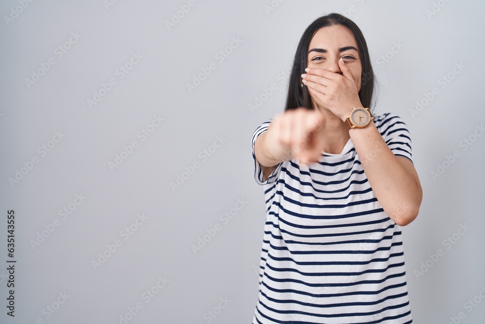 Young brunette woman wearing striped t shirt laughing at you, pointing finger to the camera with hand over mouth, shame expression Young brunette woman wearing striped t shirt laughing at you, pointing finger to the camera with hand over mouth, shame expression