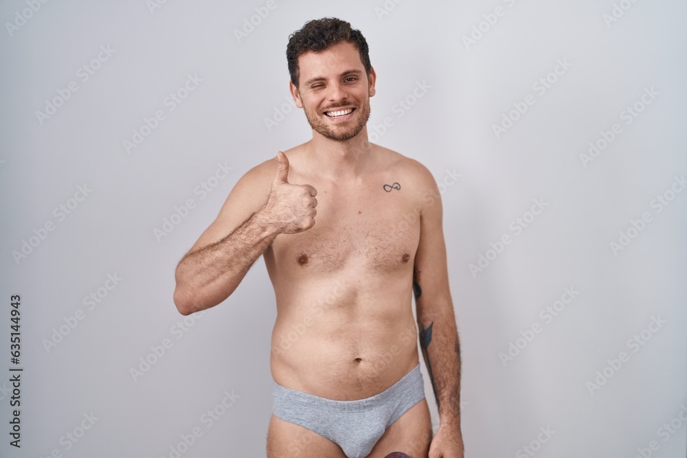 Young hispanic man standing shirtless wearing underware doing happy thumbs up gesture with hand. approving expression looking at the camera showing success.