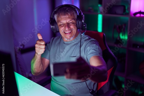 Hispanic man with grey hair playing video games with smartphone smiling happy and positive, thumb up doing excellent and approval sign