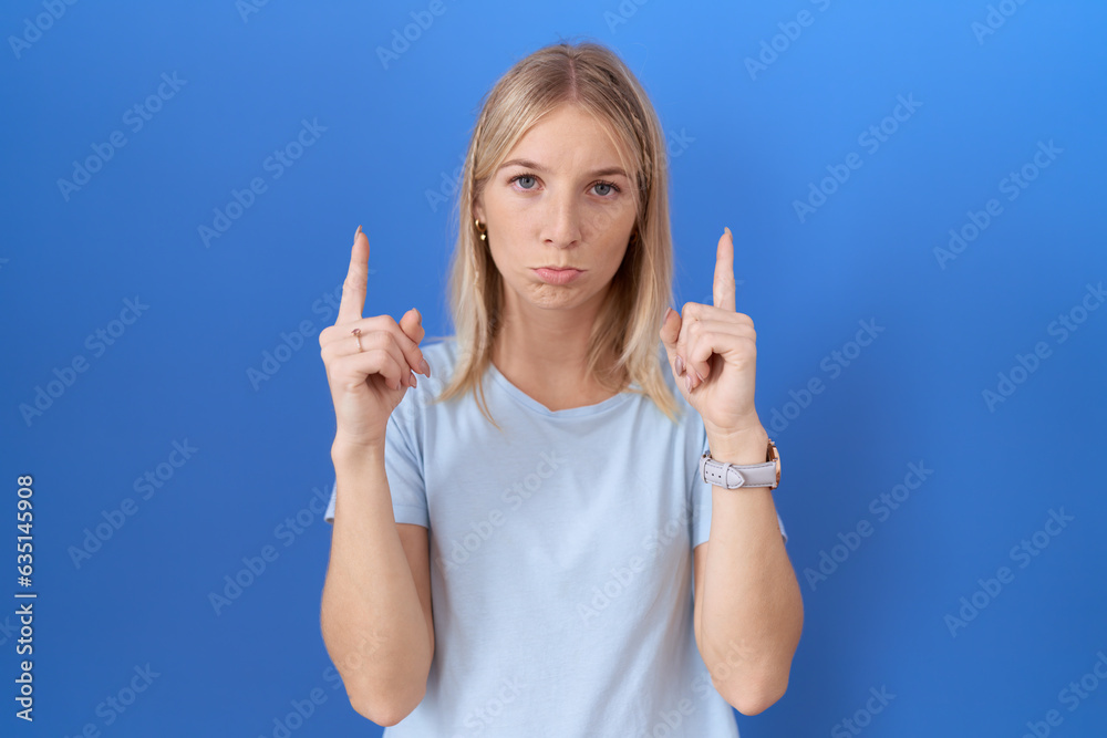 Young caucasian woman wearing casual blue t shirt pointing up looking sad and upset, indicating direction with fingers, unhappy and depressed. Young caucasian woman wearing casual blue t shirt pointing up looking sad and upset, indicating direction with fingers, unhappy and depressed.
