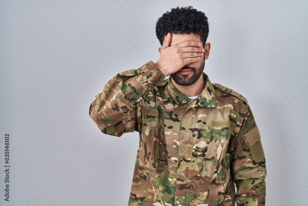 Arab man wearing camouflage army uniform smiling and laughing with hand on face covering eyes for surprise. blind concept.