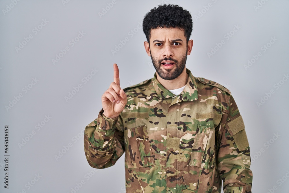 Arab man wearing camouflage army uniform pointing finger up with successful idea. exited and happy. number one.