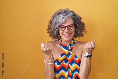 Middle age woman with grey hair standing over yellow background very happy and excited doing winner gesture with arms raised  smiling and screaming for success. celebration concept.