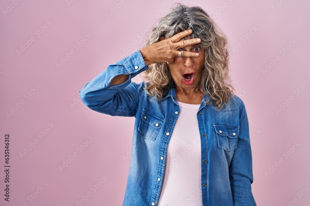 Middle age woman standing over pink background peeking in shock covering face and eyes with hand, looking through fingers with embarrassed expression. Middle age woman standing over pink background peeking in shock covering face and eyes with hand, looking through fingers with embarrassed expression.