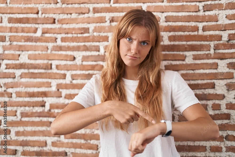 Young caucasian woman standing over bricks wall in hurry pointing to watch time, impatience, upset and angry for deadline delay Young caucasian woman standing over bricks wall in hurry pointing to watch time, impatience, upset and angry for deadline delay