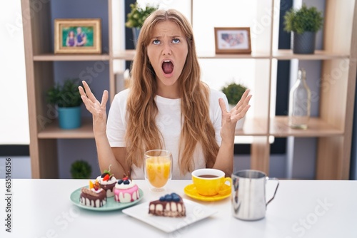 Young caucasian woman eating pastries t for breakfast crazy and mad shouting and yelling with aggressive expression and arms raised. frustration concept.