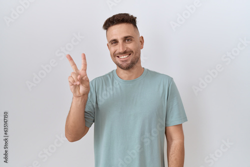 Hispanic man with beard standing over white background showing and pointing up with fingers number two while smiling confident and happy.