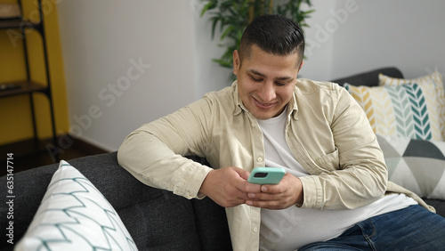 Young hispanic man using smartphone sitting on sofa at home photo