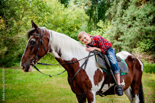 little handsome blonde smiling boy in red checkered shirt riding horse in green forest on sunny day