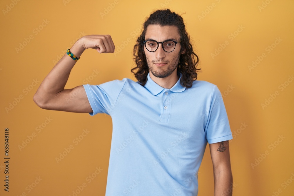 Young hispanic man standing over yellow background strong person showing arm muscle, confident and proud of power Young hispanic man standing over yellow background strong person showing arm muscle, confident and proud of power