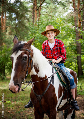 little handsome blonde smiling boy in red checkered shirt and cowboy brown hat riding horse in green forest on sunny day 