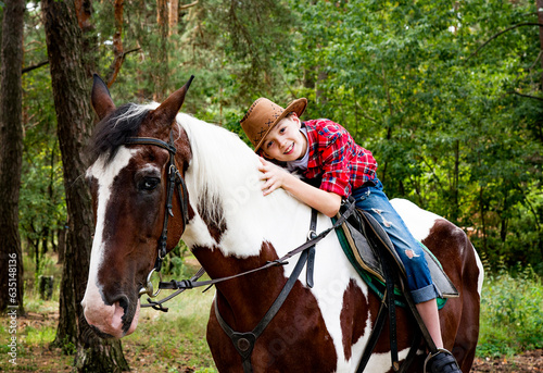 little handsome blonde smiling boy in red checkered shirt and cowboy brown hat riding horse in green forest on sunny day 