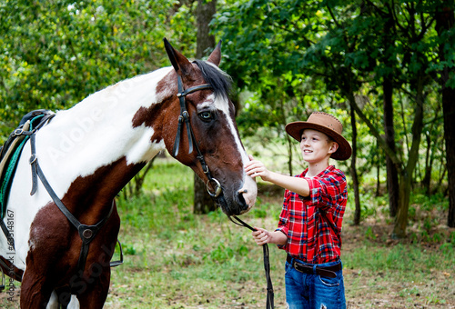 little handsome blonde smiling boy in red checkered shirt and cowboy brown hat walking and  hugging horse in green forest on sunny day  © Tetatet